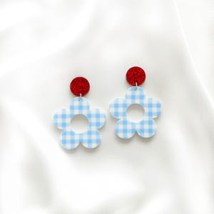 May include: A pair of flower-shaped earrings. The earrings have light blue and white gingham flower petals, with a red glittery circle at the top. The earrings are designed to dangle.