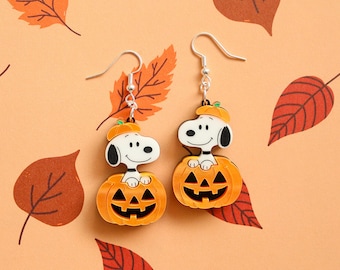 May include: A pair of dangle earrings featuring the cartoon character Snoopy wearing a small orange pumpkin hat and sitting inside a carved orange jack-o'-lantern pumpkin. The earrings have silver-toned metal hooks for attachment.