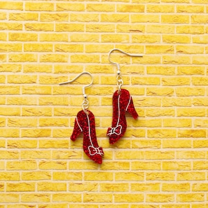 May include: A pair of red glittery high heel shoe earrings with a white bow design. The earrings hang from silver-colored fish hook ear wires. The background is a yellow brick wall.