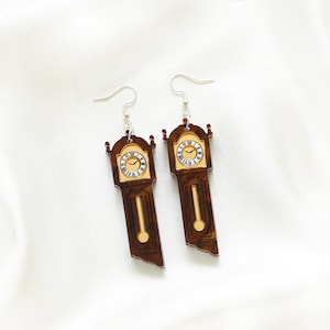 May include: A pair of dangle earrings shaped like miniature grandfather clocks. The earrings feature a brown wooden finish with gold accents and a clock face. The clock face has black Roman numerals and gold hands. The earrings hang from silver-colored fish hook ear wires.