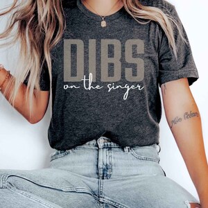May include: A dark grey t-shirt with the text "DIBS on the singer" in a light brown colour. The shirt is being worn by a person sitting on a wooden chair.