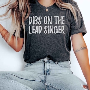 May include: A dark grey t-shirt with the text "Dibs on the Lead Singer" printed in white. The shirt is being worn by a person sitting on a wooden chair.