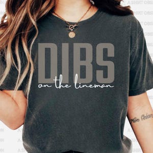 May include: Dark grey t-shirt featuring the text "DIBS on the lineman" in a bold, grey font. The phrase "on the lineman" is written in a cursive, white font. The shirt is a casual, unisex style.