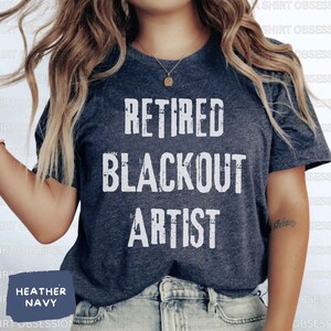 May include: A heather navy blue t-shirt with the text "Retired Blackout Artist" printed in white. The text has a distressed, vintage look.