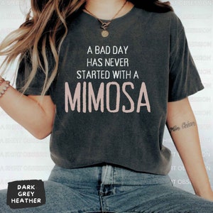 May include: A dark heather grey t-shirt with the text "A bad day has never started with a mimosa" printed in white. The word "mimosa" is in pink.