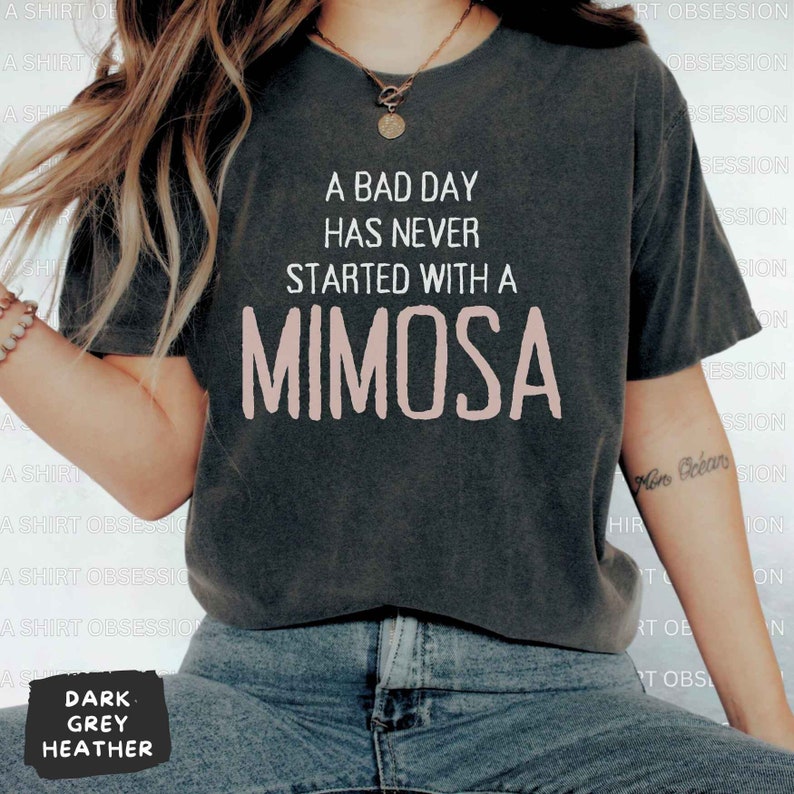 May include: A dark heather grey t-shirt with the text "A bad day has never started with a mimosa" printed in white. The word "mimosa" is in pink.