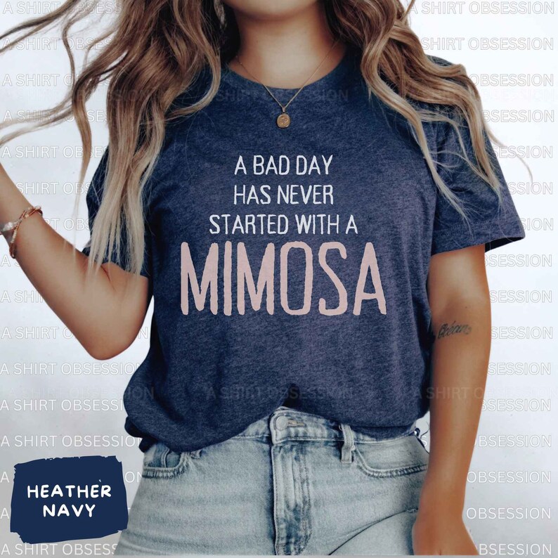 May include: A navy blue t-shirt with the text "A bad day has never started with a mimosa" in white lettering. The word "mimosa" is in pink lettering. The t-shirt is being worn by a person with long brown hair.