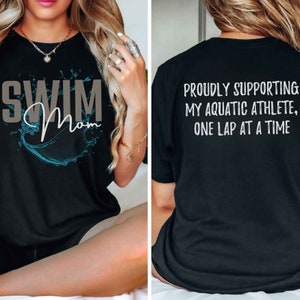 Może przedstawiać: Czarny T-shirt z napisem "Swim Mom" w kolorze szarym i niebieskim wzorem rozbryzgującej się wody z przodu. Z tyłu T-shirtu znajduje się napis "Proudly supporting my aquatic athlete, one lap at a time" w kolorze białym.