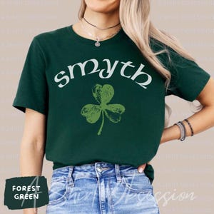 May include: A forest green t-shirt with a white graphic of a shamrock and the word "smyth" in a white cursive font.