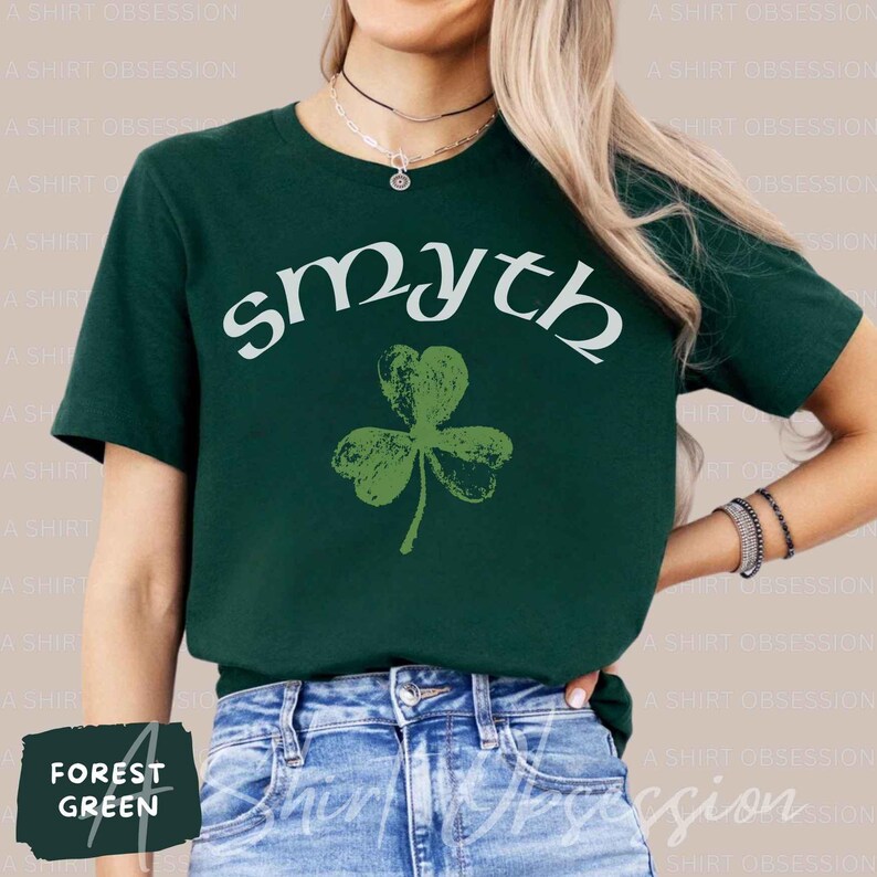 May include: A forest green t-shirt with a white graphic of a shamrock and the word "smyth" in a white cursive font.