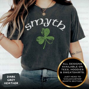 May include: A dark heather grey t-shirt with the word "smyth" in white lettering and a green shamrock graphic. The text "ALL DESIGNS AVAILABLE ON TEES, HOODIES & SWEATSHIRTS. JUST HEAD TO 'CHILD/CUSTOM' SHIRTS SECTION!" is printed on the bottom right corner of the shirt.