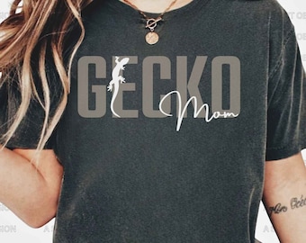 May include: A dark grey t-shirt with the text "Gecko Mom" in white lettering. A white silhouette of a gecko is to the left of the text.