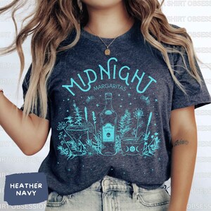 May include: A heather navy blue t-shirt with a teal blue graphic design of a bottle of tequila, a margarita glass, and a mortar and pestle. The text "Midnight Margaritas" is printed in teal blue on the shirt.