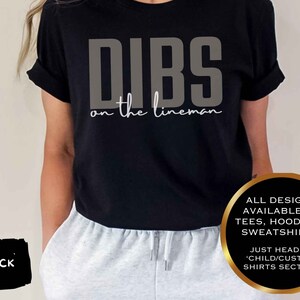 May include: Black t-shirt with the text "DIBS on the lineman" in grey and white. The shirt is worn with grey sweatpants. The image also states that all designs are available on tees, hoodies, and sweatshirts.
