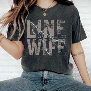 May include: A gray cropped t-shirt with the text "LINE WIFE" in a distressed font with white lightning bolts. The shirt is being worn by a person with a tattoo on their arm.