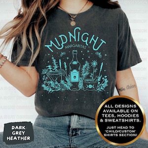 May include: A dark grey heather t-shirt with a teal design of a bottle of tequila, a margarita glass, and a mortar and pestle. The text "Midnight Margaritas" is above the design. The text "All designs available on tees, hoodies & sweatshirts. Just head to 'child/custom' shirts section!" is in the bottom right corner.