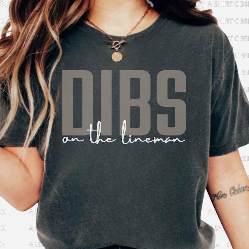May include: Dark grey t-shirt featuring the text "DIBS on the lineman" in a bold, grey font. The phrase "on the lineman" is written in a cursive, white font. The shirt is a casual, unisex style.