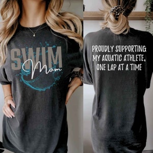 Może przedstawiać: Czarny T-shirt z niebieskim wzorem rozpryskującej się wody i napisem "Swim Mom". Z tyłu koszulki widnieje napis "Proudly supporting my aquatic athlete, one lap at a time".