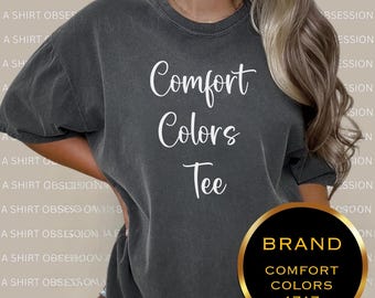 Peut inclure: Un t-shirt gris avec le texte "Comfort Colors Tee" en blanc. Le t-shirt est porté par une personne. Le nom de la marque "Comfort Colors 1717" est visible dans un cercle doré.