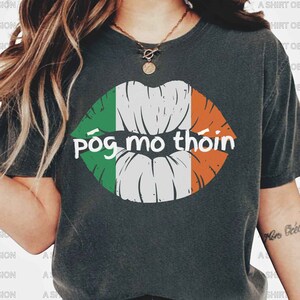 May include: A gray t-shirt with a white, green, and orange lip design and the text "póg mo thóin" printed on the front.