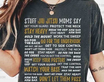 Può includere: Una maglietta grigia scura con il testo "Stuff Jiu Jitsu Moms Say" in giallo. Il testo è in una colonna verticale e include frasi come "Stay Heavy", "Go For The Back", "Watch Your Base" e "Don't Let Them Pass".