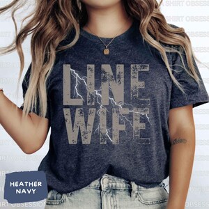 May include: A heather navy blue t-shirt with a distressed gray graphic that says "Line Wife" with a lightning bolt design.