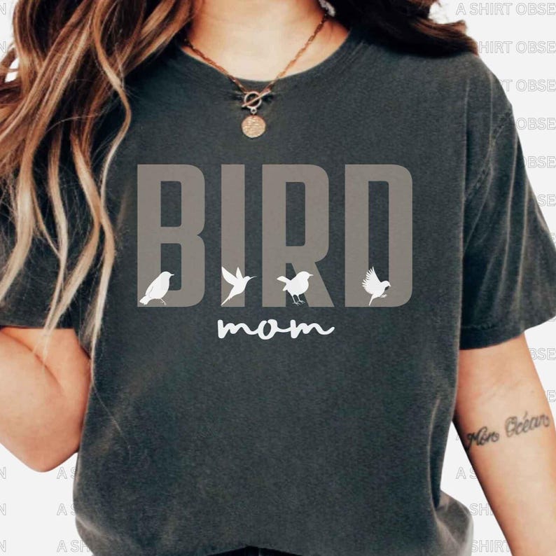 May include: A dark gray t-shirt with the word "BIRD" in white letters and a line of small white birds underneath. The text "mom" is written in cursive below the birds.