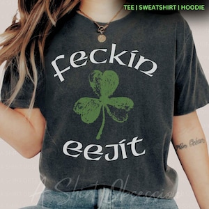 May include: A dark grey t-shirt with the text "feckin eejit" in white lettering. A green shamrock is centred above the text.