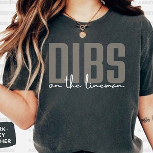 May include: Dark grey heather t-shirt with the text "DIBS on the lineman" in a bold, grey font. The phrase "on the lineman" is in a cursive, white font. The shirt is a casual style.