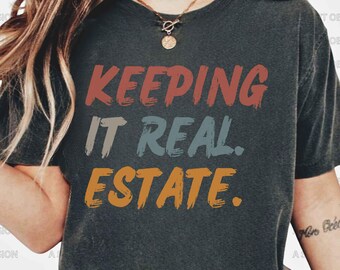 May include: A dark gray t-shirt with the text "KEEPING IT REAL. ESTATE." in a retro style font with colorful accents.