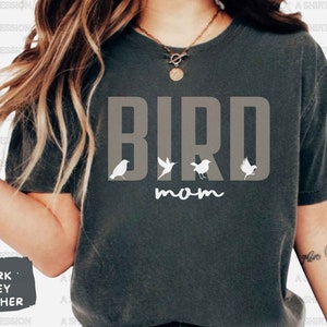 May include: Dark heather grey t-shirt with the text "BIRD mom" in white. The word "BIRD" is in large, bold letters with four small white bird silhouettes in between the letters.