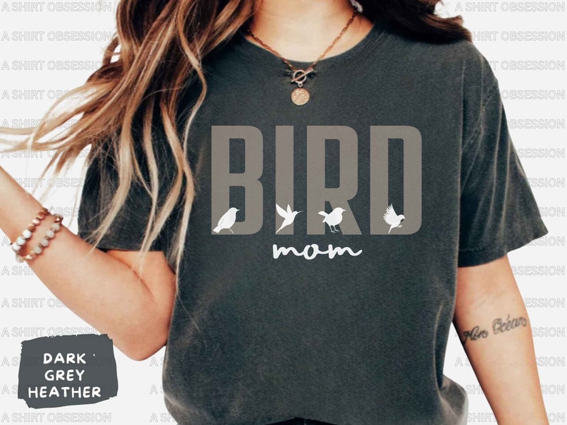 May include: Dark heather grey t-shirt with the text "BIRD mom" in white. The word "BIRD" is in large, bold letters with four small white bird silhouettes in between the letters.