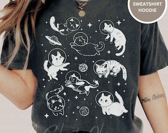 May include: Dark gray t-shirt featuring a whimsical design of white cats in space helmets, surrounded by planets and stars. The text "T-SHIRT SWEATSHIRT HOODIE" is in a circular label. A fun, space-themed graphic tee.