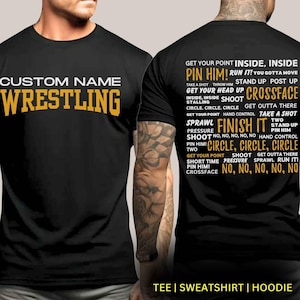May include: Black t-shirt with the text "CUSTOM NAME WRESTLING" in yellow. The back of the shirt has a list of wrestling moves in yellow text, including "PIN HIM!" "RUN IT!" "GET YOUR HEAD UP" "CROSSFACE" "SPRAWL" "FINISH IT" "SHOOT" "CIRCLE, CIRCLE, CIRCLE" and "NO, NO, NO, NO, NO".