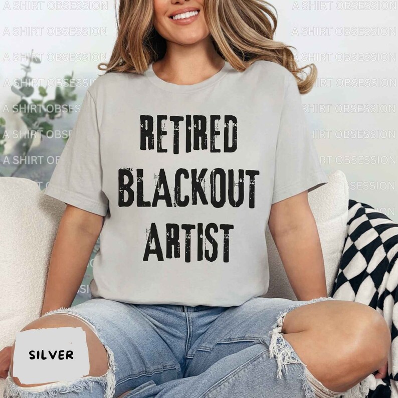May include: A gray t-shirt with the text "Retired Blackout Artist" printed in black. The shirt is being worn by a person sitting on a chair.