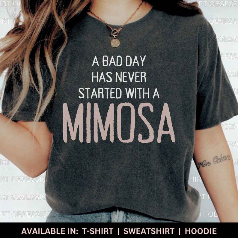 May include: A gray t-shirt with the text "A bad day has never started with a mimosa" printed in white. The word "mimosa" is in pink. The t-shirt is available in t-shirt, sweatshirt, and hoodie styles.