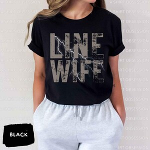 May include: Black t-shirt with the text "LINE WIFE" in gray with a lightning bolt graphic. The shirt is being worn by a person with gray sweatpants.