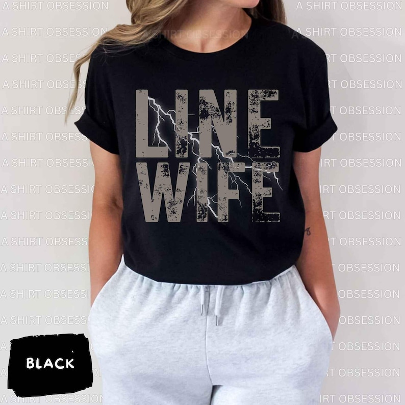 May include: Black t-shirt with the text "LINE WIFE" in gray with a lightning bolt graphic. The shirt is being worn by a person with gray sweatpants.