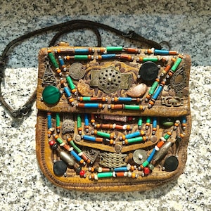 Könnte beinhalten: Eine handgefertigte, braune Leder-Umhängetasche, verziert mit bunten Perlen, Metallverzierungen und einem gewebten Riemen. Die Tasche hat einen Klappenverschluss und ist mit grünen, blauen, orangefarbenen und silbernen Akzenten verziert. Die Tasche ist etwa 20 cm breit.