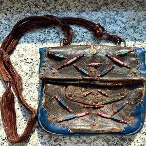 Könnte beinhalten: Vintage Leder-Umhängetasche mit braunem geflochtenem Riemen. Die Tasche hat einen Körper aus braunem Leder mit blauen Verzierungen und ist mit bunten Perlen verziert. Die Tasche hat einen Klappverschluss und eine kleine Vordertasche.