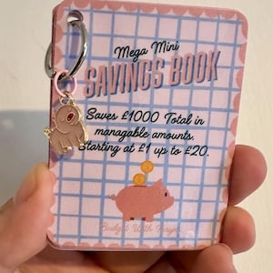 May include: A pink and blue mini savings book with the text "Mega Mini SAVINGS BOOK". The book has a pink pig charm and a silver hoop. The book's design includes a grid pattern and the text "Saves £1000 Total in manageable amounts. Starting at £1 up to £20."