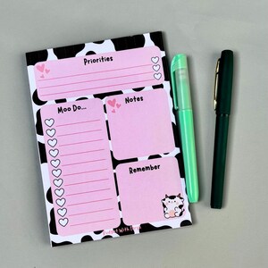 May include: A notepad with a black and white cow print border and pink sections labeled "Priorities", "Moo Do...", "Notes", and "Remember". A cartoon cow graphic is in the "Remember" section. Two pens, one green and one dark green, are next to the notepad.