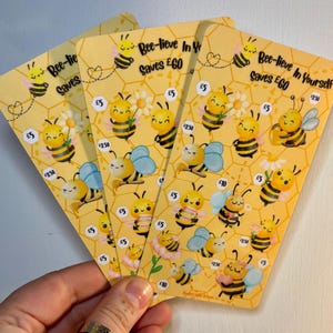 Könnte beinhalten: Drei gelbe Karten mit Illustrationen von Cartoon-Bienen und Wabenmustern. Die Karten tragen die Aufschrift "Bee-lieve In Yourself Saves £60" und zeigen Bienen und Geldbeträge.