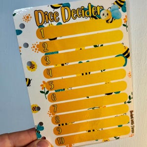 May include: A white Dice Decider card with a bee theme. The card has ten numbered slots in yellow, with bee and flower illustrations. The text "Dice Decider" is at the top. The card is held by a hand.