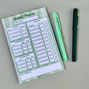 Op de afbeelding: Een groen-witte budgetplanner met madeliefjesaccenten, samen met een lichtgroene marker en een donkergroene pen. De planner bevat secties voor inkomsten, uitgaven, schuldaflossing en spaardoelen. De tekst "Budget Planner" staat bovenaan.