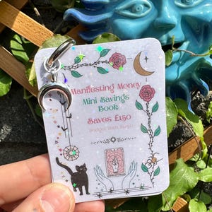 May include: A small card with the text "Manifesting Money Mini Savings Book" and a crescent moon charm. The card features illustrations of roses, a black cat, and a spider. The card is held in front of a blue sun-shaped sculpture.