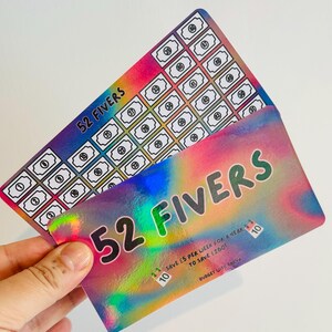 以下が含まれることがあります： 「52 Fivers」の文字が入った2枚のカラフルなカード。1枚のカードには番号付きのボックスのグリッドがあり、もう1枚には「1年間毎週5ポンド節約して260ポンド節約」と書かれています。カードは虹色のホログラフィック効果があります。