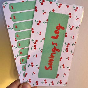 Puede incluir: Un registro de ahorros verde y blanco con un patrón de cerezas. El registro tiene ocho líneas numeradas para registrar las cantidades de ahorros. El título "Savings Log" está escrito en rojo sobre un fondo verde.