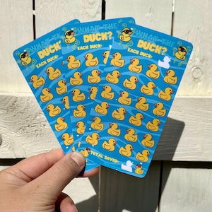Könnte beinhalten: Drei blaue Karten mit gelben Gummienten-Illustrationen und dem Text "WHAT THE DUCK?" werden gezeigt. Jede Karte hat ein Raster von Enten und die Worte "EACH DUCK". Die Karten werden vor einem weißen Holzgrund gehalten.