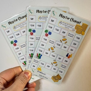 Puede incluir: Cuatro tarjetas blancas laminadas con el título "Play For Change!" y un tablero de juego con diferentes denominaciones de moneda británica. El tablero de juego tiene un punto de inicio y un punto final y los jugadores tiran un dado para determinar cuánto dinero pueden ahorrar.